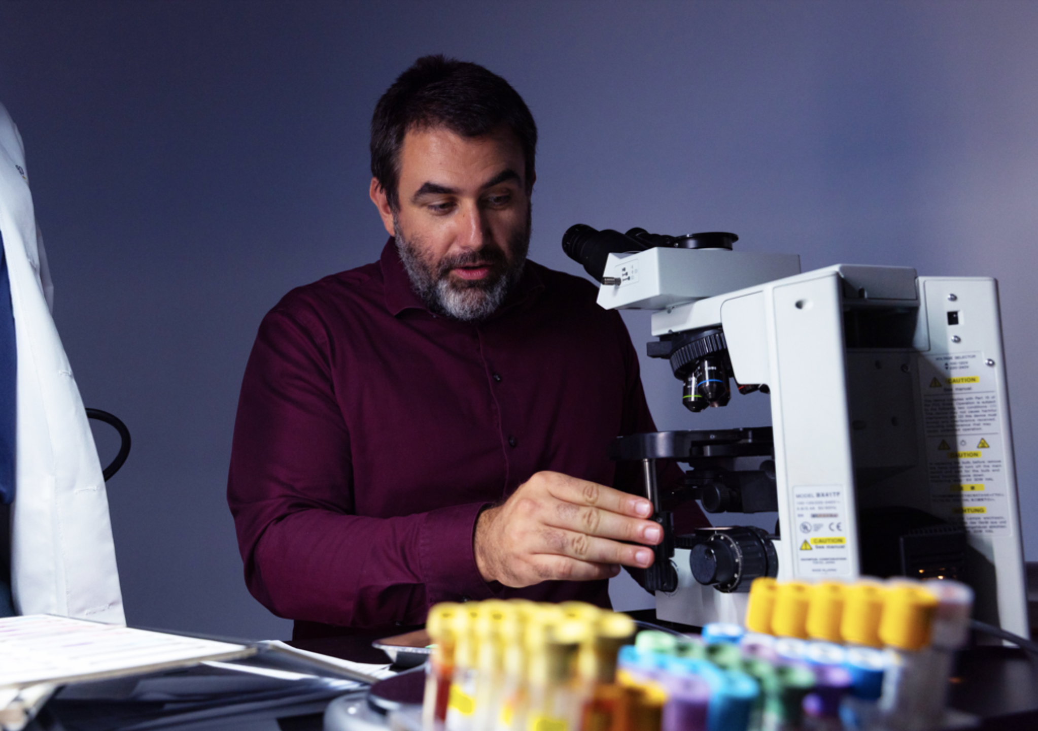 UC San Diego professor Ludmil Alexandrov adjusts a white compound microscope at a lab bench; yellow-capped test tubes are in the foreground.